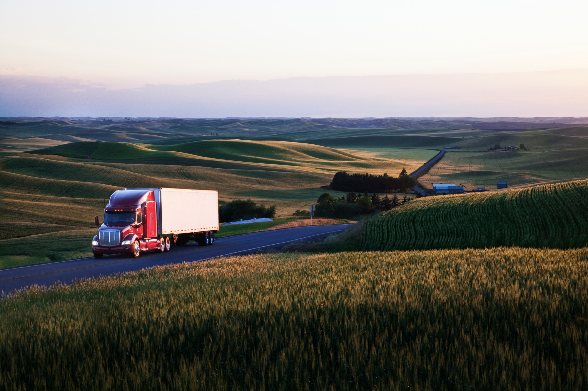 Red commercial truck driving through lush wheat fields at sunset, representing commercial vehicle operations in rural landscapes.